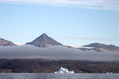 Band of mist across Arab Peaks on Danmark  with an iceberg by the shore. Scoresbysund Fiord. East Greenland. 2005